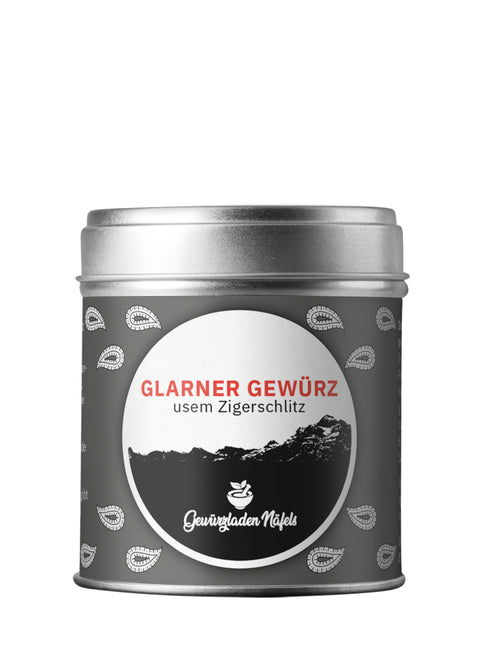 Eine silberne, zylindrische Gewürzdose für „Glarner Gewürz“ vom Gewürzladen Näfels mit einem dunkelgrauen Etikett, weißen Paisley-Mustern und einer zentralen Berg-Illustration.
