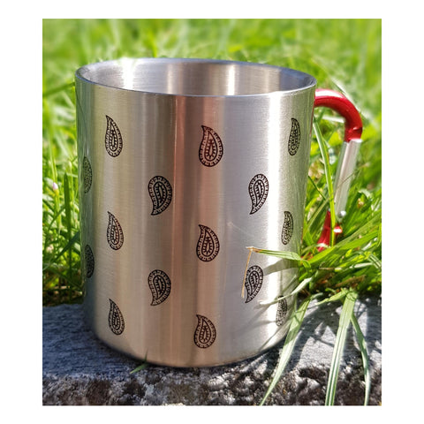 Eine Nahaufnahme eines Campingbechers aus Edelstahl mit eingraviertem schwarzem Paisley-Muster, der im Freien auf einem Stein vor einem verschwommenen grünen Grashintergrund steht.