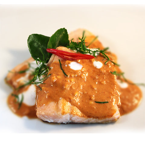 Nahaufnahme von gebratenen Lachsfilets mit dicker Panang-Currysoße, garniert mit Kaffir-Limettenblättern, Kokoscreme-Tropfen und einer aufgeschnittenen roten Chili.