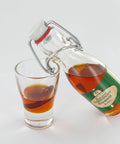 Eine klare Bügelflasche, die rötlich-orangefarbenes Peperoncini-Kräuteröl in ein kleines Glas gießt, wobei sowohl in der Flasche als auch im Glas eine ganze getrocknete Chilischote zu sehen ist.