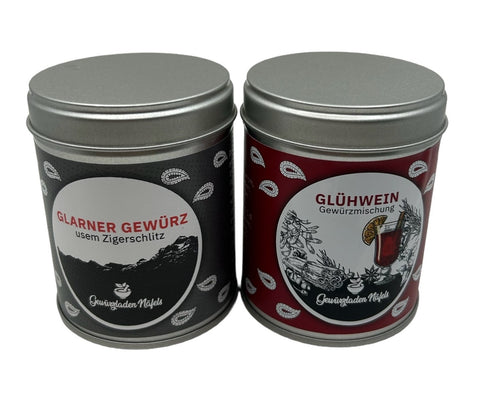 Zwei Gewürzdosen stehen nebeneinander: links eine schwarze Dose "Glarner Gewürz usem Zigerschlitz" und rechts eine rot-schwarze Dose "Glühwein Gewürzmischung".