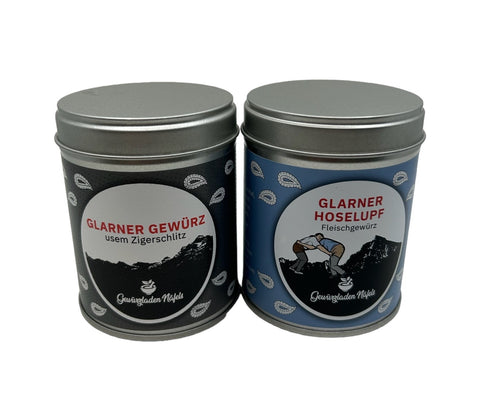 Zwei Gewürzdosen stehen nebeneinander: links eine schwarze Dose "Glarner Gewürz usem Zigerschlitz" und rechts eine blaue Dose "Glarner Hoselupf Fleischgewürz".
