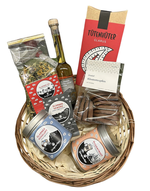 Ein Weidenkorb gefüllt mit verschiedenen Glarner Produkten aus der Schweiz, darunter eine Flasche Likör, Glarner Tee im Beutel, eine rote Schachtel Tütenhüter, eine Packung Dinkel Klostertropfen Kekse und zwei runde Dosen.