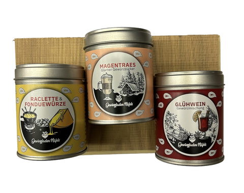 Drei etikettierte Gewürzdosen für Raclette, Glühwein und eine spezielle Mischung, präsentiert auf einem Holzpodest.