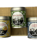 Drei bunte Gewürzdosen mit Abbildungen für Raclette, Salzkräuter und Fleischgewürz, auf einem kleinen Holzständer.