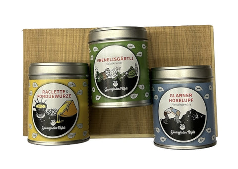 Drei bunte Gewürzdosen mit Abbildungen für Raclette, Salzkräuter und Fleischgewürz, auf einem kleinen Holzständer.