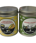 Zwei Gewürzdosen stehen nebeneinander: links eine gelbe Dose "Raclette & Fonduewürze" und rechts eine grüne Dose "Vrenelisgärtli Salzkräuter".
