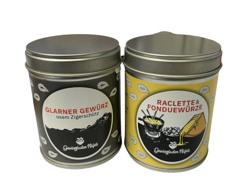Zwei Gewürzdosen stehen nebeneinander: links eine schwarze Dose "Glarner Gewürz usem Zigerschlitz" und rechts eine gelbe Dose "Raclette & Fonduewürze".