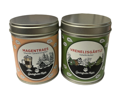 Zwei Gewürzdosen stehen nebeneinander: links eine orangefarbene Dose "Magentraes Glarner Gewürzzucker" und rechts eine grüne Dose "Vrenelisgärtli Salzkräuter".