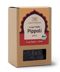 Eine braune Kartonverpackung von „Classic Ayurveda“ Pippali (Langer Pfeffer) mit einem rot-beigen Etikett, Bio-Siegel und einem kleinen Sichtfenster, das ganze schwarze Pfefferkörner zeigt.