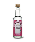 Eine klare Glasflasche „Classic Ayurveda Rosenwasser“ mit einem rosa-weißen Etikett mit Rosen-Illustrationen, goldenem Deckel und Bio-Siegel.