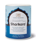 Eine zylindrische blau-weiße Dose mit „Sharkara“ ayurvedischem Rohrzucker von Classic Ayurveda, mit dekorativem Bogen-Logo und mehrsprachigen Produktbeschreibungen.
