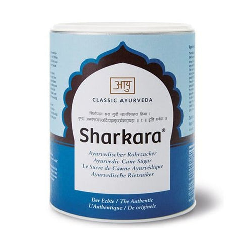 Eine zylindrische blau-weiße Dose mit „Sharkara“ ayurvedischem Rohrzucker von Classic Ayurveda, mit dekorativem Bogen-Logo und mehrsprachigen Produktbeschreibungen.