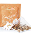 Ein orangefarbenes Tee-Sachet für Sirocco „Camomile Orange Blossoms“ Kräutertee neben einem transparenten Pyramidenteebeutel, der mit getrockneten Kamillenblüten und feinen Orangenblüten gefüllt ist.