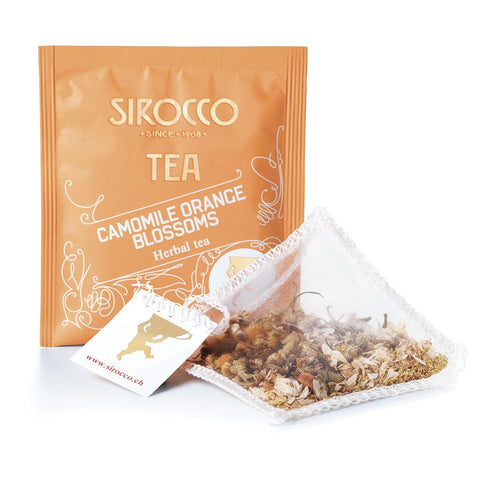 Ein orangefarbenes Tee-Sachet für Sirocco „Camomile Orange Blossoms“ Kräutertee neben einem transparenten Pyramidenteebeutel, der mit getrockneten Kamillenblüten und feinen Orangenblüten gefüllt ist.