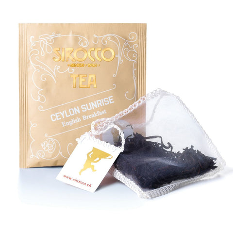 Ein beigefarbenes Tee-Sachet für Sirocco „Ceylon Sunrise“ English Breakfast Tee, hinter einem transparenten Pyramidenteebeutel, der mit dunklem, langblättrigem Schwarztee gefüllt ist.