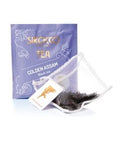 Ein lavendelfarbenes Tee-Sachet für Sirocco „Golden Assam“ Schwarztee, platziert hinter einem transparenten Pyramidenteebeutel, der mit feinen, dunklen Schwarzteeblättern gefüllt ist.