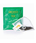 Ein smaragdgrünes Tee-Sachet für Sirocco „Green Jasmine“-Tee hinter einem transparenten Pyramidenteebeutel, der mit dunklen, duftenden Grünteeblättern gefüllt ist.