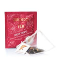 Ein dunkelrotes Tee-Sachet für Sirocco „Green Tropic“ Grüntee mit goldener Aufschrift und einem transparenten Pyramidenteebeutel, der mit Grünteeblättern und exotischen Fruchtstücken gefüllt ist.