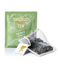 Ein olivgrünes Tee-Sachet für Sirocco „Piz Palü“ Kräutertee, abgebildet mit einem transparenten Pyramidenteebeutel, der eine Bergkräutermischung aus getrockneten grünen Blättern und Blüten enthält.