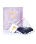 Ein zartviolettes Tee-Sachet für Sirocco „Purple Breeze“ Darjeeling-Tee hinter einem transparenten Pyramidenteebeutel, der mit dunklen, hochwertigen Schwarzteeblättern gefüllt ist.