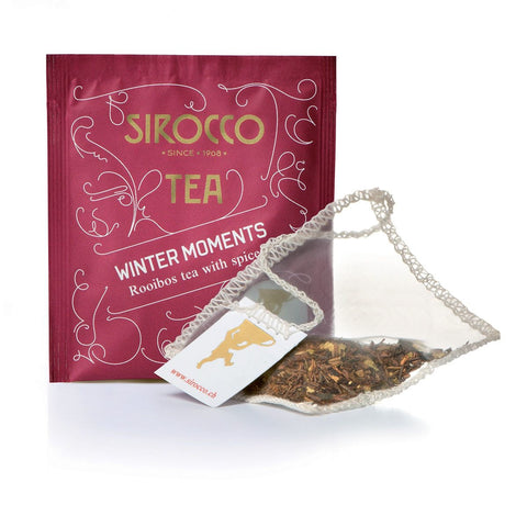 Ein tiefrotes Tee-Sachet für Sirocco „Winter Moments“ neben einem transparenten Pyramidenteebeutel, der mit rötlich-braunen Rooibos-Teeblättern und feinen Gewürzstücken gefüllt ist.