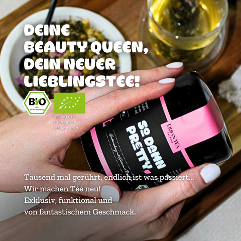 Eine Hand hält die schwarze Dose Urban Tea Factory "So Damn Pretty" Grünteemischung mit rosa Band. Im Hintergrund sind eine Schale mit dem losen Tee und eine Tasse des aufgebrühten Tees zu sehen. Das BIO-Siegel ist ebenfalls abgebildet.