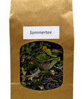 Eine braune Papiertüte „Sommertee“ mit einem ovalen Sichtfenster, das eine lose Kräuterteemischung aus grünen Blättern und bunten Blütenblättern zeigt.