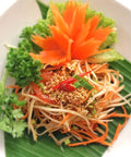 Eine Portion Som Tum (Thailändischer Grüner Papayasalat), präsentiert auf einem Bananenblatt in einem weißen Gericht, garniert mit Salat, Koriander, Erdnüssen, Chili und einer geschnitzten Karottenblume.