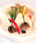 Nahaufnahme einer weißen Schüssel mit cremiger Tom Kha Suppe, die Stücke von Huhn oder Tofu, Pilze, Zwiebelringe, Lauchzwiebeln und eine rote Chili-Scheibe enthält.