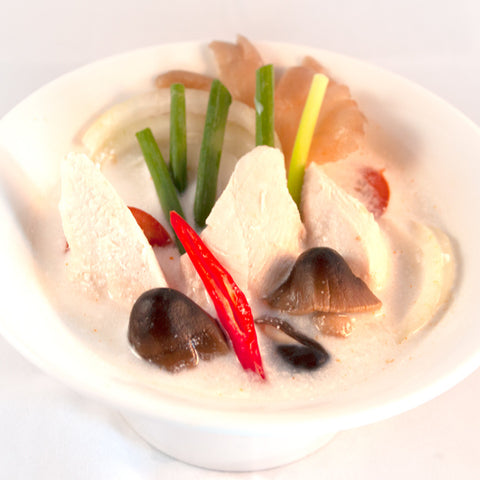 Nahaufnahme einer weißen Schüssel mit cremiger Tom Kha Suppe, die Stücke von Huhn oder Tofu, Pilze, Zwiebelringe, Lauchzwiebeln und eine rote Chili-Scheibe enthält.
