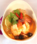 Eine Portion Tom Yum Suppe in einer weißen Schale, garniert mit zwei großen Garnelen, Limettenscheibe, Koriander, roten Chilischoten, Pilzen und einer lila Orchidee daneben.