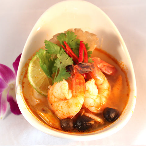 Eine Portion Tom Yum Suppe in einer weißen Schale, garniert mit zwei großen Garnelen, Limettenscheibe, Koriander, roten Chilischoten, Pilzen und einer lila Orchidee daneben.