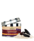 Eine silberne Metalldose Herbaria „Tonkabohnen ganz, im Zuckerbett“ mit offenem Deckel, daneben drei ganze dunkle Tonkabohnen auf einem kleinen Haufen weißem Zucker.