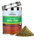 Eine silberne Gewürzdose von „Herbaria“ mit dem Etikett „Wilde Hilde“ für Salate, in grün-braunem Design mit Bio-Siegel, neben einem kleinen Haufen der groben Kräutermischung.