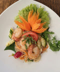 Eine Portion Yam Wun Sen (Thailändischer Glasnudelsalat) mit Garnelen, Gurke, Tomate und Koriander, kunstvoll garniert mit einer geschnitzten Karottenblume und Salat auf einem weißen, dreieckigen Teller.