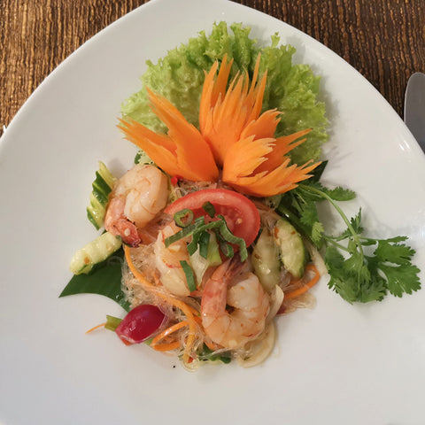 Eine Portion Yam Wun Sen (Thailändischer Glasnudelsalat) mit Garnelen, Gurke, Tomate und Koriander, kunstvoll garniert mit einer geschnitzten Karottenblume und Salat auf einem weißen, dreieckigen Teller.