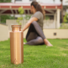 Kupfer-Yoga-Flasche im Gras, mit einer Person, die im Hintergrund eine sitzende Drehhaltung macht.